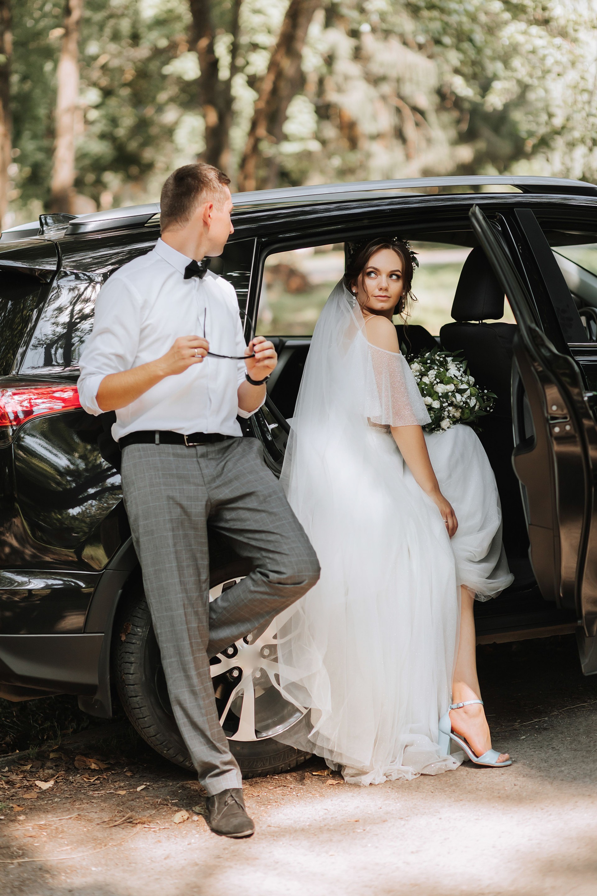 Wedding portrait, photo of a stylish groom in a white shirt and bow tie and a brunette bride with a bouquet of flowers near a black car.